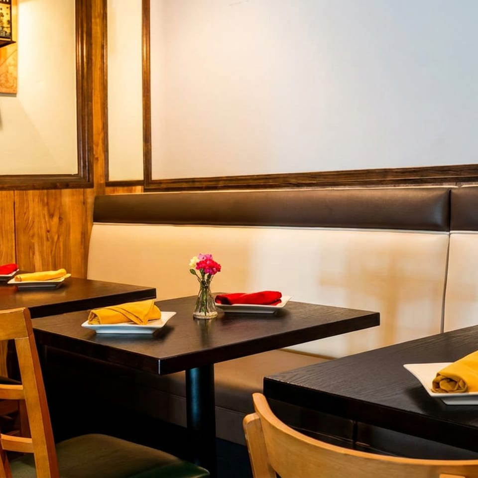 Empty restaurant dining room with dark tables, wooden chairs, and a long booth with light and dark cushions.
