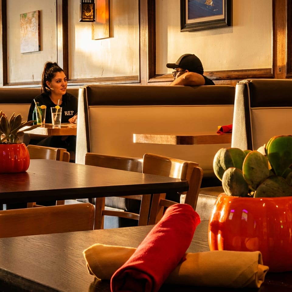 A restaurant interior with booth seating, dark tables, and potted plants, with two patrons in the background.