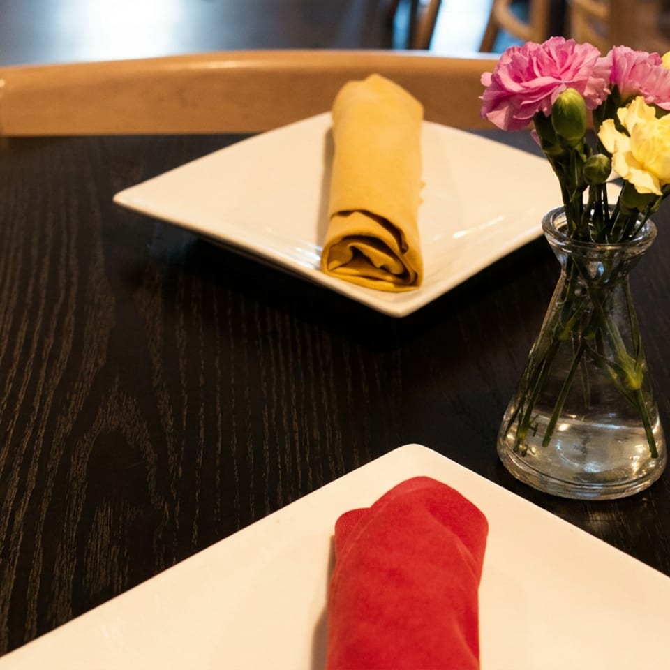 Two place settings with rolled napkins on white plates and a small vase of pink and yellow flowers.
