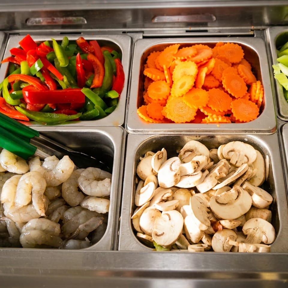 A buffet-style selection of fresh ingredients including meats, shrimp, bell peppers, carrots, mushrooms, and tomatoes.